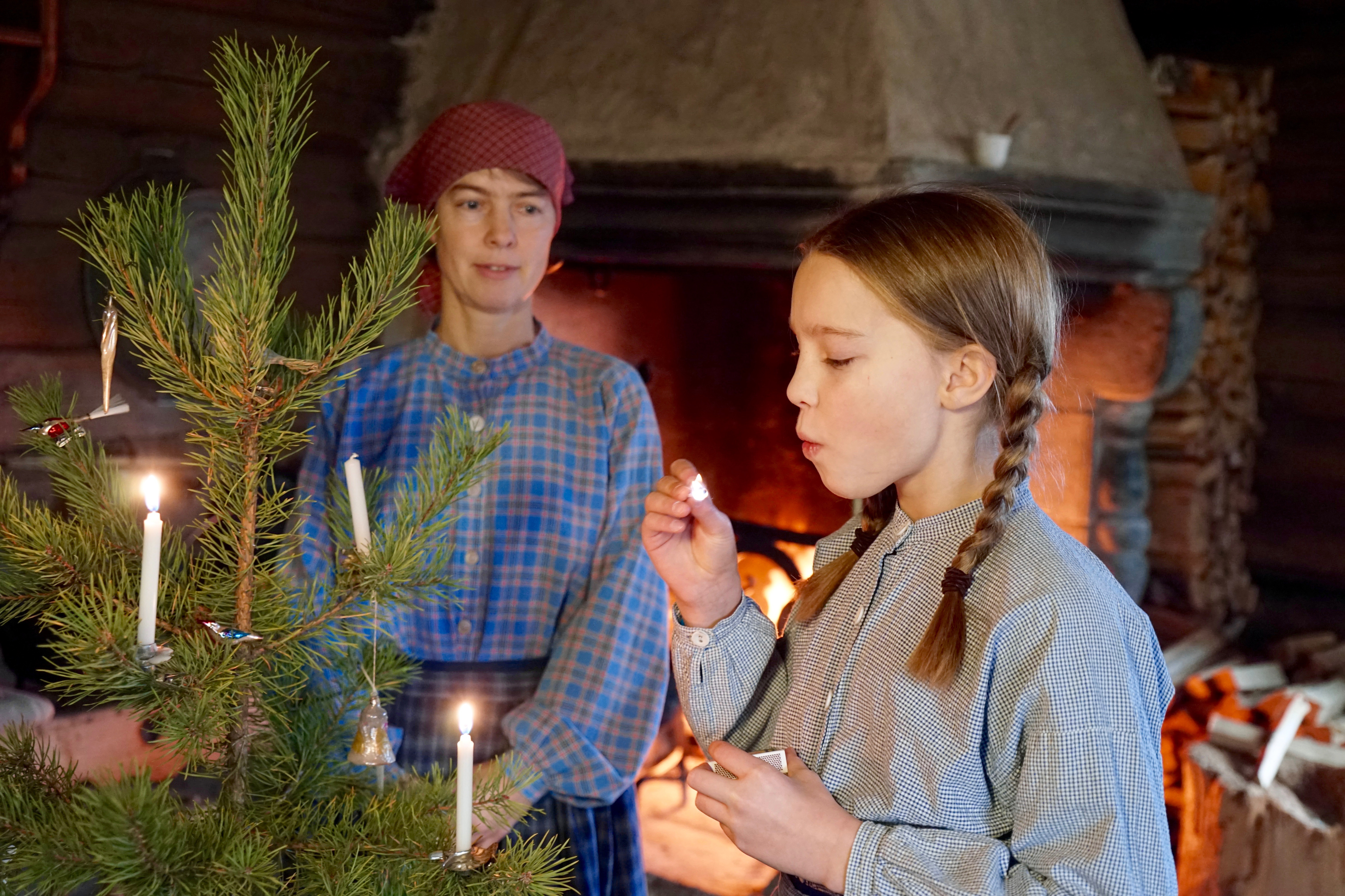 Jente i historiske klær blåser ut en fyrstikk etter å ha tent stearinlys på juletreet.