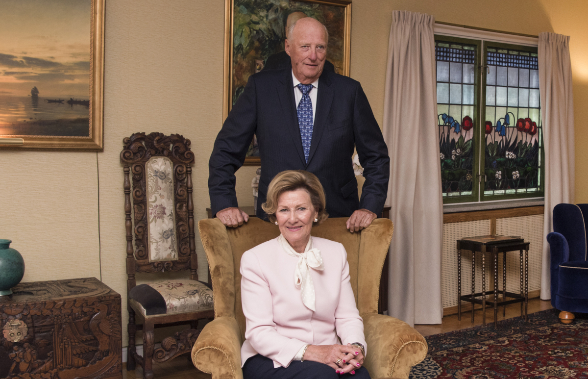 Dronning Sonja sittende i en lenestol med kong Harald stående bak i stuen i dronningens barndomshjem på Maihaugen.