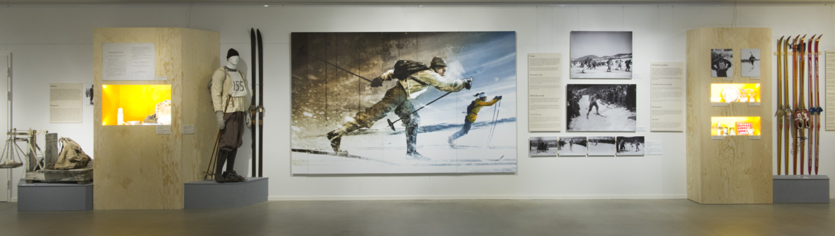 Utstillingslokale med stor illustrasjon på veggen av en kvinnelig skiløper i moderne antrekk som går fra en skiløper med anorakk og gammeldags utstyr. Også flere mindre bilder, montre og gamle ski utstilt.